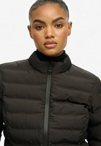 Czarna puchowa kurtka z wysokim kołnierzem, z pikowanym wypełnieniem, zamkiem z przodu i gładką teksturą. Nosi się ją na czarnym golfie.