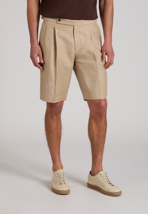 Uomo che indossa pantaloncini pieghettati beige, scarpe da ginnastica crema e una maglietta marrone, in piedi su uno sfondo chiaro uniforme.