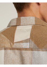 Camisa de cuadros marrón y crema con una textura de fieltro, que presenta un panel crema en contraste en la sisa y un detalle de costura visible.