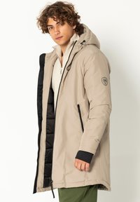 Beige Parkajacke mit schwarzem Futter, Frontreißverschluss, Kapuze und Seitentaschen. Kontrastärmel und Badge-Detail am Oberarm.