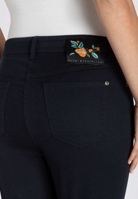 Jeans bleu foncé avec un tissu lisse, taille mi-haute avec une étiquette florale, et design classique à cinq poches avec couture décorative.