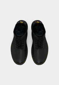 Een paar zwarte leren veterschoenen met gele stiksels en merkpulltabs, van bovenaf gezien op een witte achtergrond.