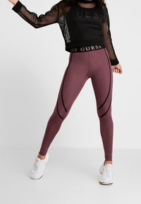 Czarna siateczkowa bluzka z długim rękawem z logo "GUESS"; bordowe legginsy z czarnymi bocznymi panelami; w połączeniu z białymi butami sportowymi.