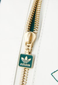 Balta medžiaga su auksine užtrauktuku, turinti stačiakampį žalią etiketę su Adidas logotipu ir auksiniu traukimo užtrauktuku. Matoma tekstūrinė siuvimo detalė.
