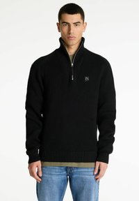 Schwarzer gerippter Pullover mit Reißverschlusskragen, versehen mit einem kleinen Logo-Patch auf der Brust. Getragen über einem hellgrünen Hemd, kombiniert mit blauer Jeans.