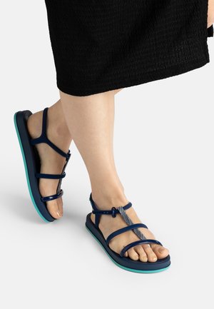 Füße mit dunkelblauen Riemchensandalen mit türkisfarbenen Sohlen, ein Fuß leicht angehoben, vor weißem Hintergrund und sichtbarem Saum eines schwarzen Rocks.