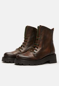 Botas de tobillo de cuero marrón con cordones y acentos verdes, superficie texturada y una suela gruesa de goma negra con un patrón de tracción robusto.