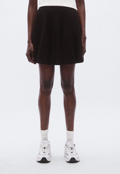 Jupe plissée noire avec une taille ajustée, longueur au-dessus du genou, associée à des baskets blanches et des chaussettes basses. Tissu lisse et texturé.