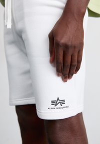 Hvide bomulds-shorts med et lille sort logo på venstre ben, glat tekstur og snøre i taljen. Nærbillede af hånd på shorts.