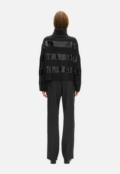 Persona che indossa un maglione a collo alto nero con strisce orizzontali lucide e pantaloni larghi neri, in piedi di spalle su uno sfondo bianco.