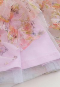 Tissu rose léger à motif floral aux couleurs douces, avec des couches de tulle transparent et des accents de volants le long des bords.