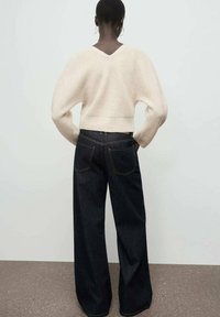 Pullover di maglia crema con un texture a coste e maniche ampie, abbinato a jeans in denim blu scuro a gamba larga con tasche posteriori e cuciture gialle.