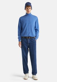 Muž oblečený v modrém roláku, tmavě modrých džínách s černým opaskem, námořnické pletené čepici a bílých teniskách, stojící s jednou rukou v kapse.