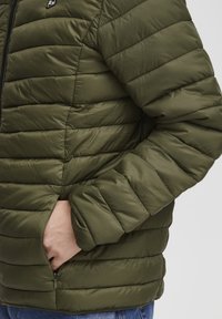 Personne portant une veste matelassée vert olive avec la main dans la poche zippée, montrant la manche et la vue latérale.