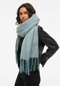 Lichtblauwe, gestructureerde sjaal van zacht materiaal met rafelige uiteinden, gedragen rond de nek over een zwarte leren jas.