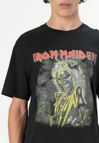 Melnas kokvilnas t-krekla, ar lielu pelēku un dzeltenu grafiku, kurā attēlots skeleta figūra, ar "Iron Maiden" tekstu sarkanā krāsā virs dizaina.