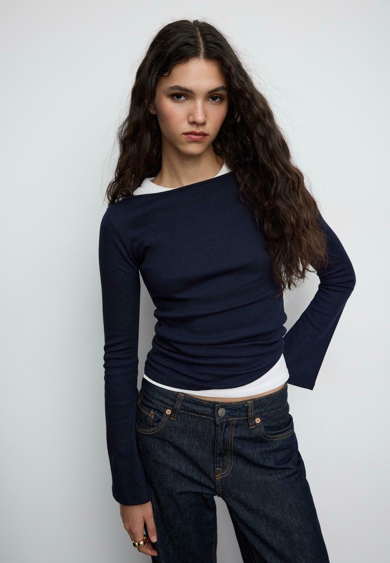 Temno modra majica z dolgimi rokavi iz rebrastega materiala, nošena preko bele srajce. Kombinirana s temnimi jeans kavbojkami z oranžnim šivom. Gladek tekstura.