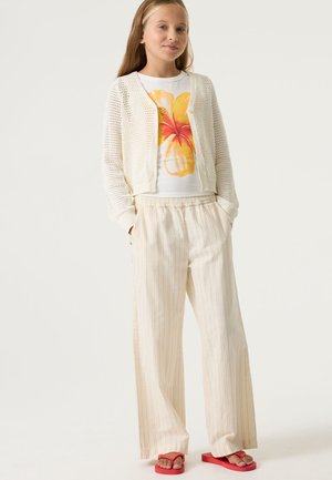 Lichtgewicht gebreid crèmekleurig vest over een wit T-shirt met een gele bloem, gecombineerd met gestreepte beige wijde broekspijpen en rode slippers.