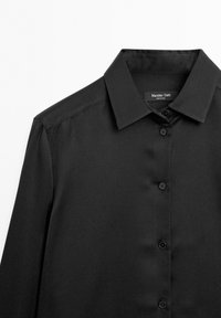 Black button-up shirt with a classic collar, smooth fabric, and long sleeves. Features a label on the inside collar. Six black buttons down the front.