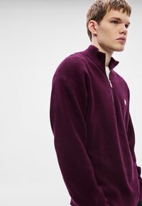 Μωβ πουλόβερ με φερμουάρ μέχρι τη μέση, κατασκευασμένο από υφασμάτινο ύφασμα με υφή, διαθέτει μακριά μανίκια και ένα μικρό έμβλημα στο στήθος, τοποθετημένο σε ουδέτερο φόντο.