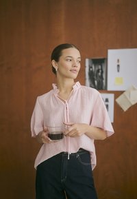 Light pink, textured blouse with a ruffled neckline, button closure, worn with dark jeans. Holding a transparent glass of coffee.