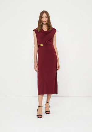 Kvinde står iført en ærmeløs burgundy midi-kjole med en guldbroche og sorte hæle med ankelrem mod en ensfarvet hvid baggrund.