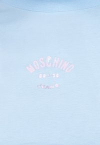 Ανοιχτό γαλάζιο ύφασμα με το κείμενο «MOSCHINO 86 • 23 JEANS» σε ανοιχτό ροζ, διατεταγμένο σε μικρό τόξο.