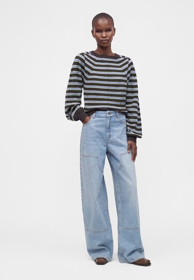 Femme portant un pull rayé noir, blanc et bleu avec un jean bleu clair à jambes larges et des chaussures marron, debout avec une main dans la poche.
