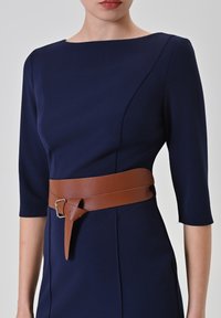Marineblaues, figurbetontes Kleid mit dreiviertel Ärmeln, das mit einem braunen Ledergürtel in der Taille gebunden ist und die Silhouette betont.