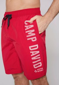 Piros úszónadrág rugalmas derékrésszel és zsinórral, az oldalán fehér felirattal: "CAMP DAVID" és "WATER SPORT". Sima textúra.