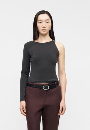 Mladá žena stojí v asymetrickom čiernom topu s jedným dlhým rukávom a jedným odhaleným plecom, v kombinácii s bordovými nohavicami a čiernym opaskom.