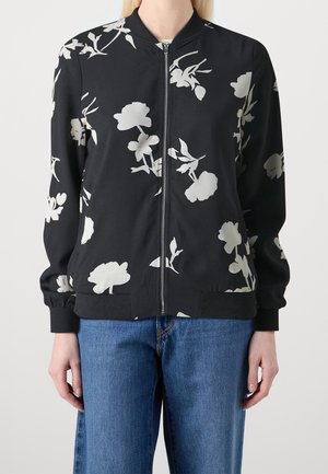 Sort lynlåstrøje med hvide blomsterprint, båret over blå jeans, vist fra nakke til hofter med hænder afslappet ved siderne.