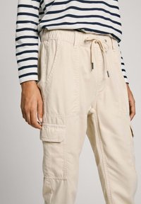 Beige cargo pants with an elastic waistband and drawstring, featuring side pockets and a flap pocket on the thigh. Smooth fabric texture.