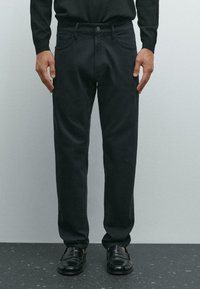 Uomo che indossa pantaloni neri a gamba dritta e mocassini di pelle neri, in piedi contro un muro grigio chiaro su un pavimento scuro a macchie.