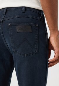Tamno plave traperice s teksturiranim završetkom. Imaju stražnji džep s crnom kožnom oznakom Wrangler i dekorativnim šavovima.
