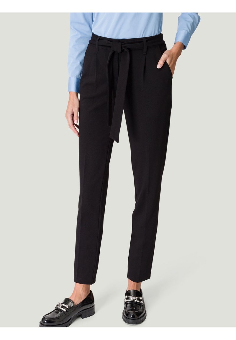 Personne portant un pantalon noir taille haute avec une ceinture nouée devant, associé à une chemise bleu clair et des mocassins noirs brillants.