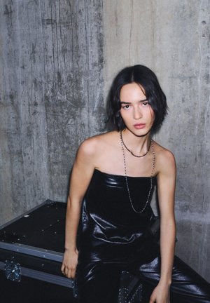 Svart, ärmlös, glansig skinn-topp kombinerad med matchande skinnbyxor. Modellen bär ett långt pärlhalsband och sitter mot en betongvägg.