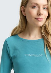 Tealfarbendes Langarm-T-Shirt aus weicher Baumwolle mit Rundhalsausschnitt und "TOM TAILOR"-Logo aus Strass auf der Brust.