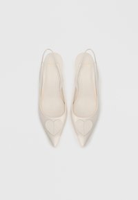 Sapatos de salto alto slingback de pele verniz bege com biqueira pontiaguda e acentos em forma de coração no peito do pé. Textura lisa e design minimalista.