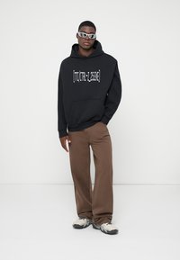 Μαύρη φούτερ με κουκούλα και λευκό κείμενο, καφέ χαλαρά παντελόνια και γκρίζα αθλητικά παπούτσια. Τα αξεσουάρ περιλαμβάνουν μοντέρνα γυαλιά ηλίου.