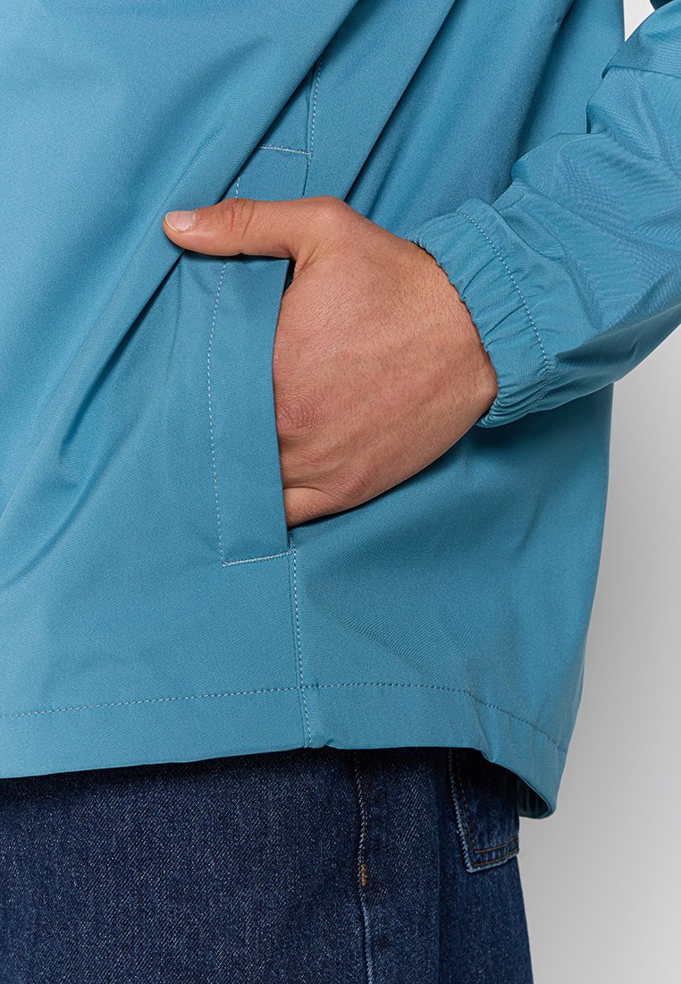 Hellblaues Windbreaker-Jacke mit glatter Textur, ausgestattet mit einem elastischen Bündchen, seitlicher Tasche und lockerer Passform.