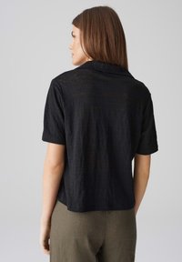 Camicia nera a maniche corte con colletto, caratterizzata da una vestibilità morbida e un tessuto testurizzato, vista da dietro.