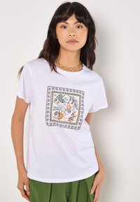 T-shirt bianco in cotone con stampa botanica quadrata nei colori verde, arancione e giallo, con bordo decorato. Maniche corte e colletto rotondo.