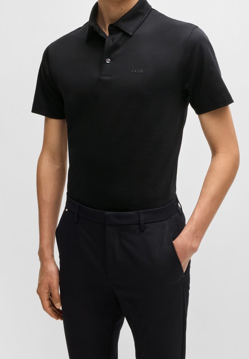 Polo noir avec col, manches courtes, patte de boutonnage et marquage subtil. Porté avec un pantalon noir ajusté, mettant en valeur un design épuré.