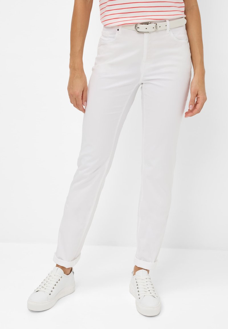 Witte katoenen broek met een rechte pijp, voorzien van een riem en opgerolde mouwen. Gecombineerd met witte sneakers, tegen een effen achtergrond.