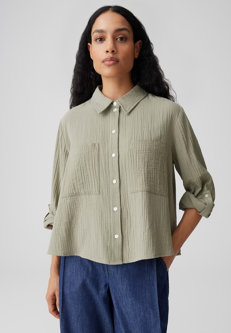 Femme aux longs cheveux bouclés portant une chemise boutonnée vert olive clair à manches retroussées et un pantalon bleu, sur un fond uni.