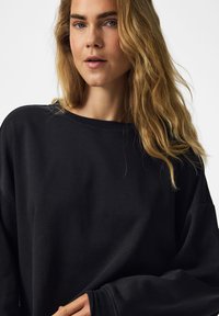 Jeune femme aux longs cheveux blonds portant un pull noir ample, regardant légèrement sur le côté avec une expression neutre.