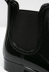 Bottine noire en cuir verni avec un panneau latéral en tissu extensible et une semelle en caoutchouc texturée. Finition lisse avec des détails de couture nets.