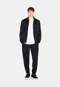 Tuta nera con piping bianco, composta da una giacca con zip e pantaloni affusolati. Indossata sopra una t-shirt bianca, abbinata a sneaker nere.