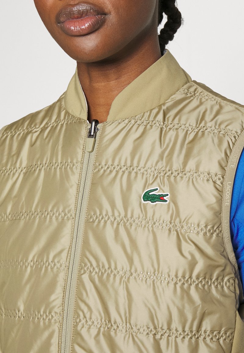 Nahaufnahme einer Person in einer beigen gesteppten Zip-Weste mit einem grünen Lacoste-Krokodil-Logo auf der Brust über einem blauen Kleidungsstück.